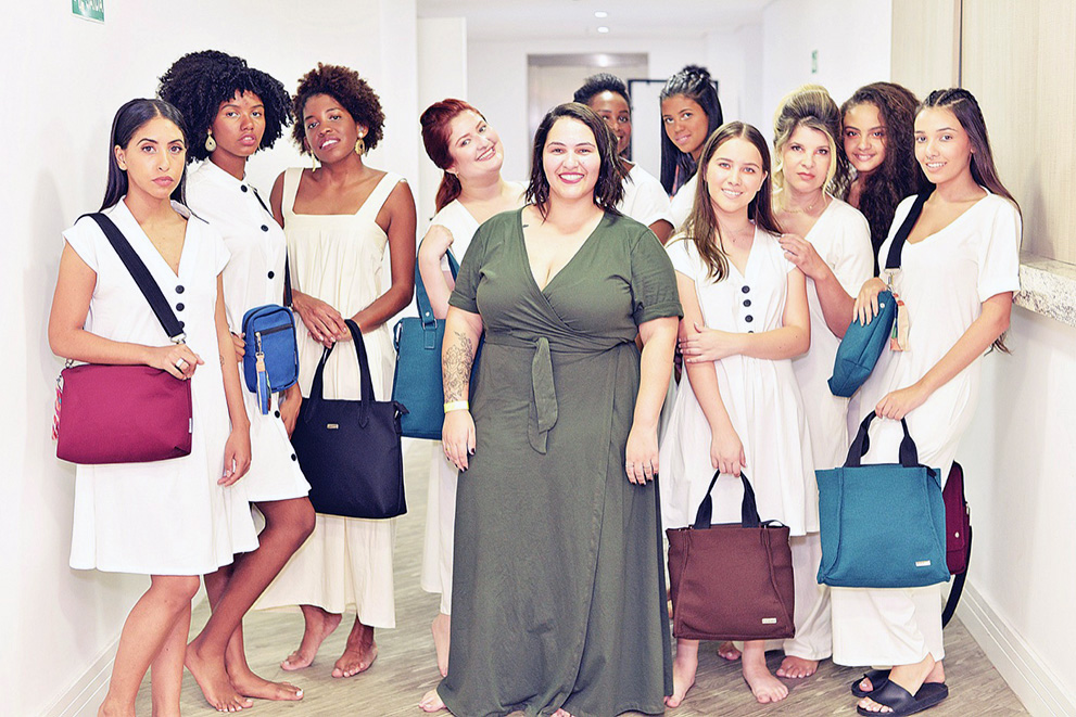 Adriana veste um vestido verde-musgo e está no centro da foto. Ao seu redor, dez mulheres vestidas de branco, todas descalças, posam com vários modelos de bolsa, de cores diferentes.