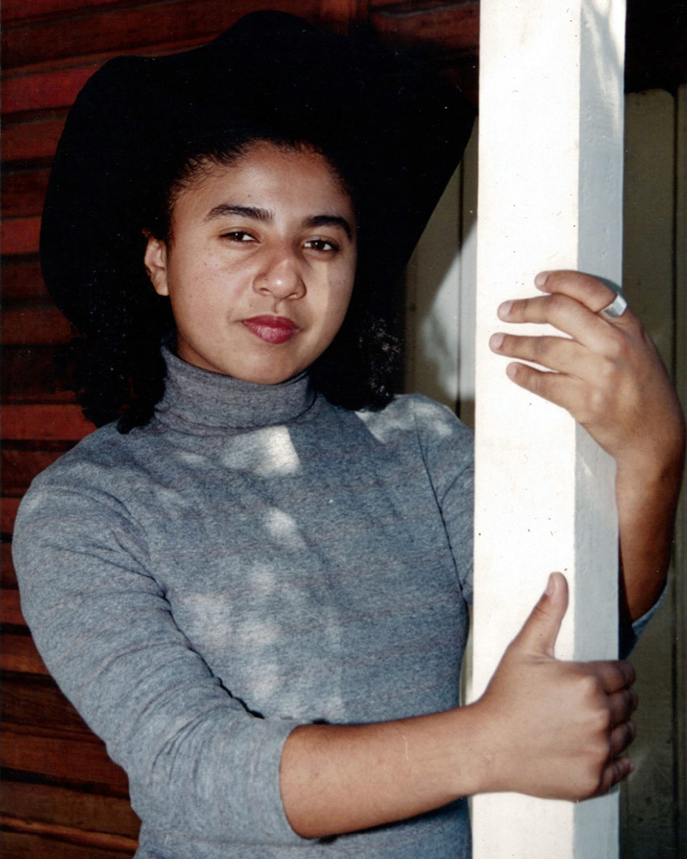 Adriana posa olhando séria para a câmera e abraçando um pilar branco. Ela usa batom, um chapéu de caubói preto e uma blusa cinza de mangas compridas.