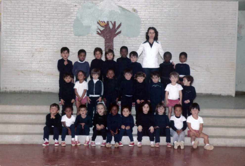 Maria Antonieta, de blazer branco, posa com uma turma de crianças. A maioria delas veste uniforme azul-marinho, algumas vestem camisetas brancas. A turma está organizada em fileiras. Ao fundo, vê-se a colagem de uma árvore na parede.
