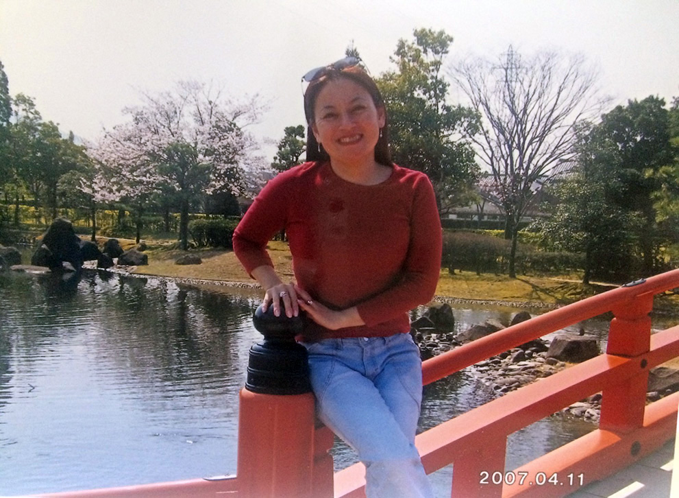Luzia está no Japão, sorrindo para a foto em uma ponte vermelha. Ao fundo, vê-se um lago calmo, pedras e algumas árvores sem folhas. 