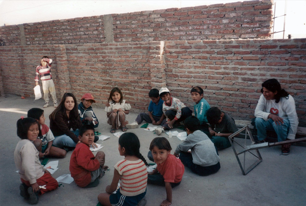 Sueli  está sentada no chão em roda com diversas crianças e papeis espalhados. Algumas olham para a câmera. Ao fundo, uma parede de tijolos. 